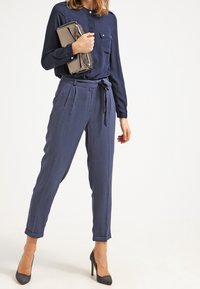 Blusa blu navy a maniche lunghe con bottone sul davanti e una tasca sul petto, abbinata a pantaloni aderenti a vita alta e tacchi grigi.