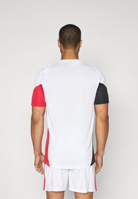 Maillot de sport à manches courtes en blanc, rouge et noir. Il présente un design avec blocs de couleur, une texture lisse et un col rond.