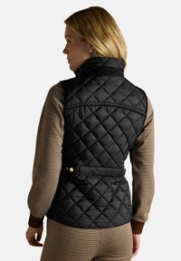 Kobieta z blond włosami, ubrana w czarną pikowaną kamizelkę bez rękawów, na brązowej bluzce w kratę z długim rękawem oraz dopasowanych spodniach, stojąca bokiem.