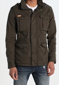 Olivengrøn lynlåstrøje med høj krave, flere forlommer og justerbar talje. Fremstillet af holdbart stof med en glat tekstur.