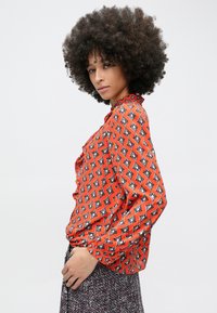 Sarkana blūze ar dimanta formas rakstu, garām piedurknēm un augstu apkakli. Audums šķiet gluds ar vieglu spīdumu.