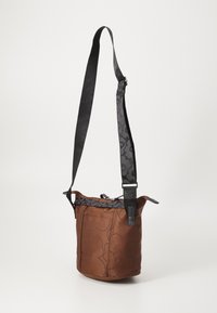 Brown fabric bucket bag with black floral pattern. Adjustable black strap, zip closure, textured surface, and side seam pockets.