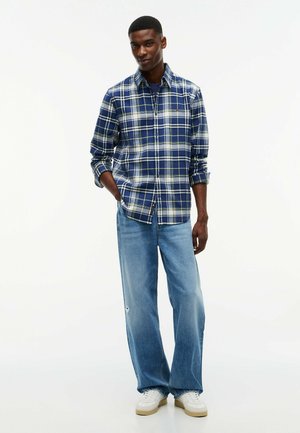 Uomo che indossa una camicia a quadri blu, jeans azzurri a gamba larga, sneakers bianche, in piedi con una mano in tasca su uno sfondo bianco semplice.
