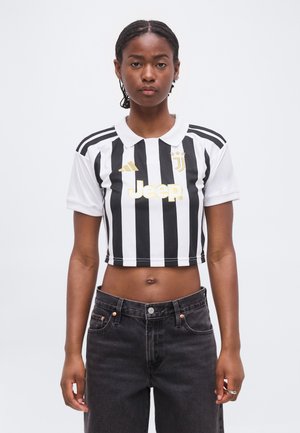 Joven mujer con trenzas, que lleva una camiseta de fútbol corta a rayas blancas y negras y vaqueros negros, de pie frente a un fondo liso.
