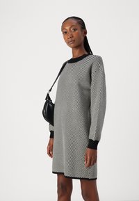 Vestido em padrão espinha de peixe preto e branco com mangas longas, gola e punhos canelados pretos, combinado com uma pequena bolsa preta. Textura suave.