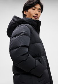 Schwarze Daunenjacke mit Kapuze, mit gesteppten Abschnitten, elastischen Bündchen und einem Reißverschluss vorne. Hergestellt aus einem leichten Stoff.
