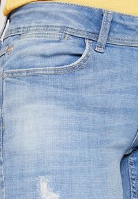 Ljust blå denimjeans med en något sliten textur, med en framficka, synliga sömmar och en liten rivning nära lårområdet.