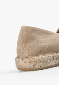 Beige canvas espadrille-schoen met een gestructureerde bovenkant, gesloten neus en een gevlochten jute zool, die een natuurlijke, aardse uitstraling heeft.