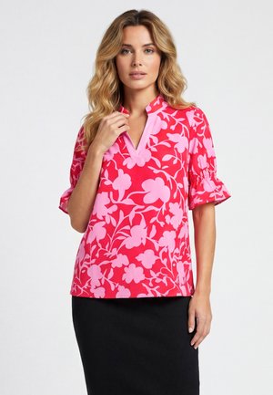 Vrouw met golvend blond haar, die een rode en roze bloes met bloemenprint en een zwarte rok draagt, poseren met één hand dicht bij het sleutelbeen.
