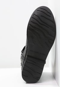 Sandale synthétique noire avec surface texturée, ferrures en argent et semelle à motifs. Comprend une sangle réglable et un logo de marque bien visible sur la semelle.