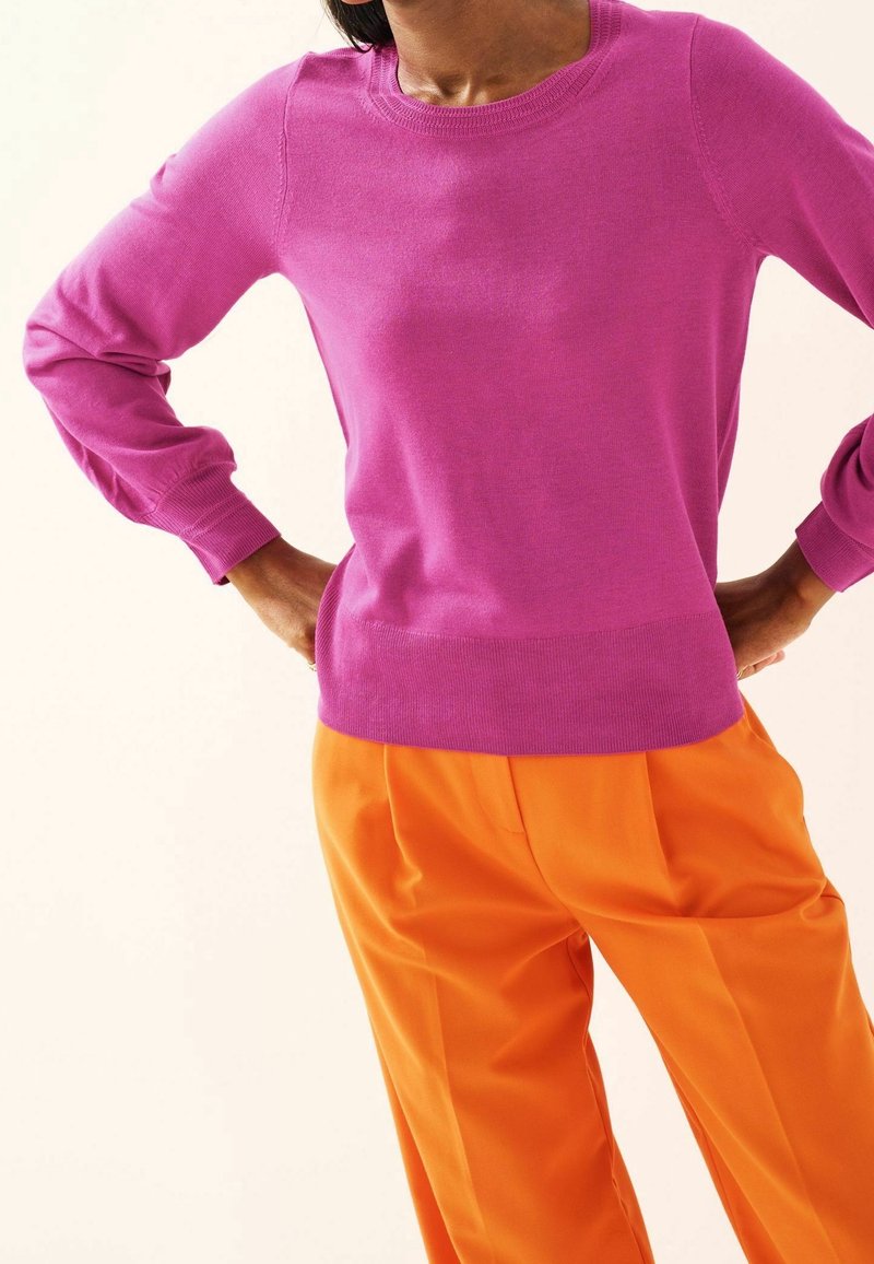 Violetti neulepaita, jossa on pitkät hiharesorit ja ribbireunukset, yhdistetty kirkkaan oransseihin laskostettuihin housuihin. Sujuva pinta, klassinen pyöreä kaula-aukko.