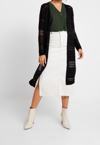 Black knitted cardigan with open weave design, green top, white denim skirt with front zipper, and cream heeled ankle boots.