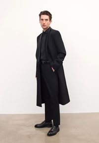 Black tailored coat, matching shirt and pants, leather shoes with a strap detail, smooth texture, minimalist design against a light backdrop.