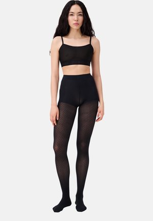 Donna con capelli lunghi e scuri che indossa un top corto nero e collant neri a vita alta con fantasia, in piedi davanti a uno sfondo bianco.