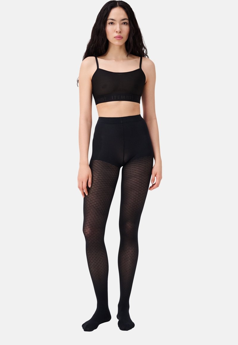 Femme aux longs cheveux noirs portant un débardeur noir court et des collants noirs à motifs taille haute, debout devant un fond blanc.