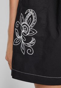 Robe en coton noire ornée de broderies florales blanches sur la poche gauche, avec de fines coutures le long de l'ourlet pour plus de détail.