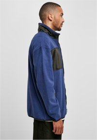 Giacca in pile blu con una tasca sul petto in nylon nero e colletto, caratterizzata da una zip completa e una vestibilità rilassata. Il tessuto ha una consistenza morbida.