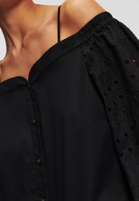 Svart blus med off-shoulder-design, med en texturerad, broderad ärm och knappstängning. Slät tyg med veckade detaljer.