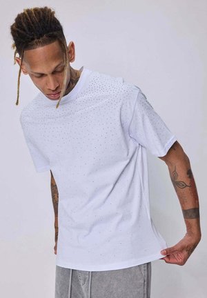 Homme avec des tatouages et des dreadlocks portant une chemise blanche à manches courtes avec de petits points, tirant sur l'ourlet de la chemise, regardant vers le bas.