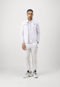Balts sporta uzvalks ar sarkaniem un zilajiem akcentiem, ar zip-up jaku un atbilstošām biksēm. Valkāts ar baltām sporta apaviem.