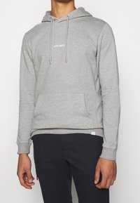 Sudadera gris hecha de una mezcla de algodón, con un bolsillo frontal, capucha ajustable y logotipo blanco "LES DEUX" en el pecho. Textura suave, corte regular.