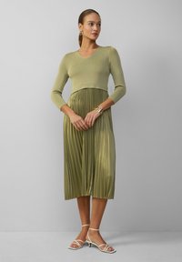 Abito verde oliva con parte superiore aderente in maglia a coste, gonna plissettata in satin e sandali bianchi con cinturini. Bracciale dorato come accessorio.