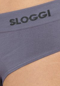 Lilla stoftrusse med en glat tekstur. Har et sort elastisk bånd med "SLOGGI"-logo. Side sømmen er synlig nær kanten.