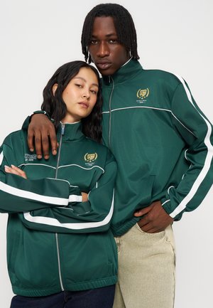 Deux personnes portant des vestes de survêtement vertes assorties avec des bandes blanches posent proches l'une de l'autre, l'une reposant son bras sur l'épaule de l'autre.