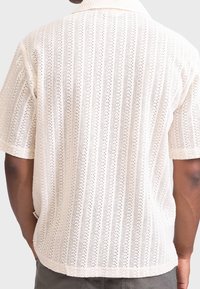 White short-sleeve shirt featuring a textured, open-knit design with vertical patterns and a fold-down collar. Hem is slightly curved.