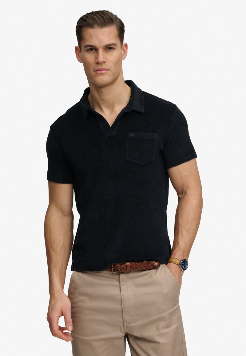 Man draagt een zwart polo met korte mouwen en borstzak, beige broek, gevlochten bruine riem en horloge, staand tegen een witte achtergrond.