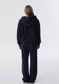 Persoon met lang haar, gekleed in een marineblauwe hoodie en bijpassende losse broek, staande met de rug naar de camera tegen een effen achtergrond.