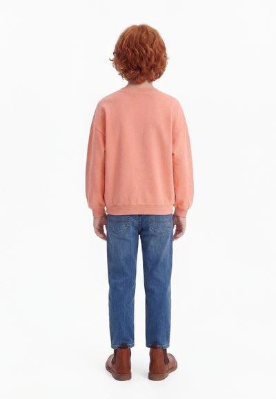 Enfant aux cheveux roux bouclés portant un sweat-shirt pêche, un jean bleu et des bottes marron, debout de dos sur un fond blanc.