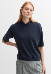 Felpa corta a maniche lunghe color blu navy con vestibilità rilassata, dotata di collo con cordino, texture liscia e vita elasticizzata. Pantaloni a quadretti grigi chiari.