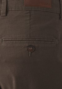Gros plan sur un pantalon en tissu brun texturé, mettant en avant une poche arrière avec une ouverture horizontale, accentuée par un petit bouton marron et une étiquette en cuir.