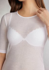 Haut blanc côtelé à manches courtes présentant une texture transparente et le contour visible d'un soutien-gorge blanc en dessous. Encolure ronde et design ajusté.