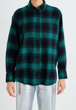 Personne portant une chemise à carreaux verte et noire boutonnée et un pantalon argenté métallique sur un fond uni clair.