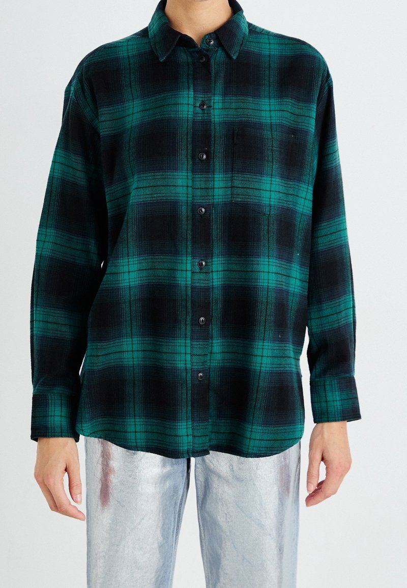 Personne portant une chemise à carreaux verte et noire boutonnée et un pantalon argenté métallique sur un fond uni clair.