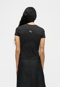 Γυναίκα με μακριά σκούρα μαλλιά που φοράει μαύρο κοντομάνικο τοπ Puma και μαύρη φούστα, γυρισμένη με την πλάτη σε απλό ανοιχτό φόντο.