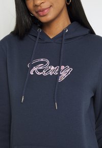 Sweat à capuche bleu marine en coton avec cordons de serrage. Présente un logo "Roxy" rose brodé. Texture lisse avec des accents de ferrures argentées.