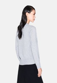 Vrouw met lang, donker haar draagt een lichtgrijze gebreide trui en een zwarte rok, poseert en kijkt naar rechts tegen een effen achtergrond.