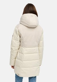Woman wearing a cream-colored, padded winter coat with a large hood, standing facing away against a plain light background.