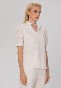 Blouse blanche en broderie anglaise avec un col en V, des manches courtes bouffantes et un col accentué. Présente des perforations rondes sur tout le tissu.