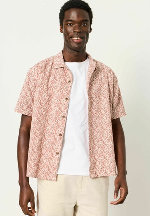 Lächelnder Mann, der ein kurzärmeliges, hellrotes, gemustertes Hemd über einem weißen T-Shirt und beigefarbene Hose trägt, steht vor einem schlichten Hintergrund.