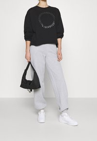 Svart sweatshirt med ett cirkulärt grafiskt mönster, grå sweatpants och vita sneakers. Håller en svart tygväska med ett silverlås.