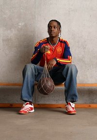 Κόκκινο και μπλε αθλητικό σετ με κίτρινες λεπτομέρειες; μπλε τζιν; κρατώντας μια καφέ μπάλα μπάσκετ σε μία νετ. Λευκά και κόκκινα αθλητικά παπούτσια. Τσιμεντένιο φόντο.