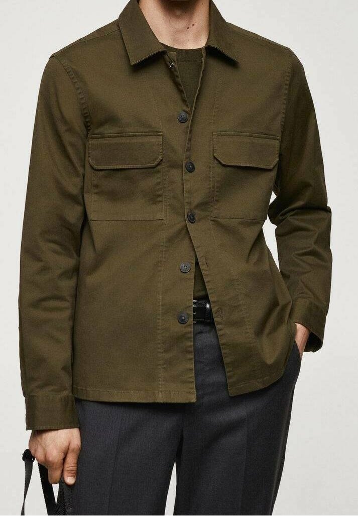 Homme portant une veste-chemise vert olive avec des poches poitrine et un pantalon foncé, une main dans la poche et tenant une sangle.