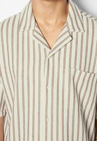 Koszula z krótkim rękawem z kołnierzykiem w jasnym kremowym kolorze z pionowymi paskami w odcieniach brązu i stonowanego różu. Posiada naszywaną kieszonkę i fakturowaną tkaninę.