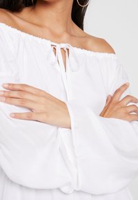 Blouse blanche à épaules dénudées avec un nœud à l'avant, des manches cloche longues et une coupe décontractée, fabriquée en tissu léger.