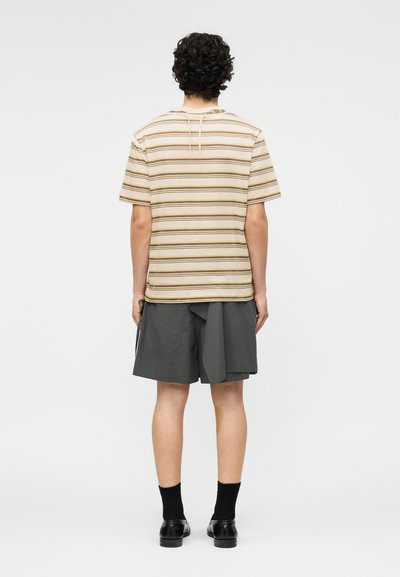 Personne aux cheveux bouclés vue de dos, portant une chemise à manches courtes rayée beige et marron, un short gris, des chaussettes noires et des chaussures noires.