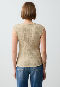 Beige mouwloos topje met een gestructureerd, gekreukeld oppervlak, een nauwsluitende pasvorm en zijsplitten; gecombineerd met hoogwaisted blauwe spijkerbroek.